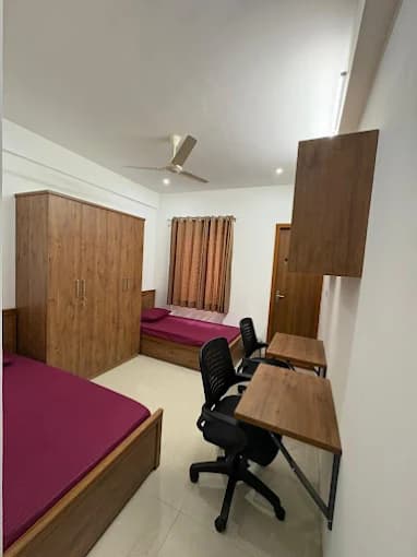 Small bedroom with two purple beds, a wooden wardrobe, and two desks with black chairs.