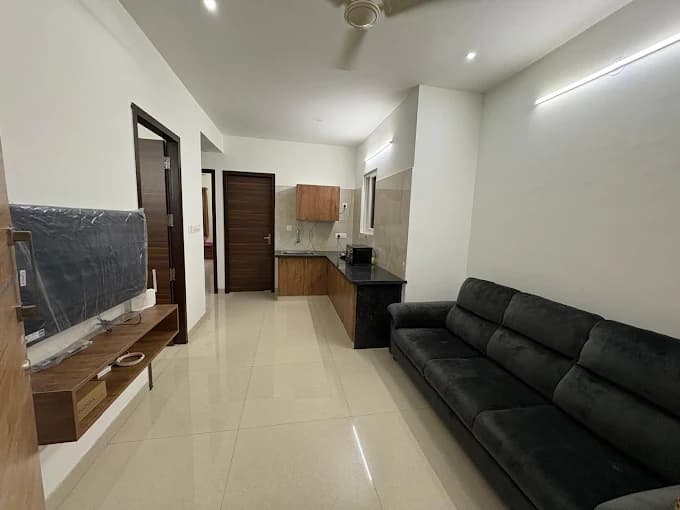 Modern living space featuring a long black sofa, wall-mounted TV, and small wooden kitchenette.