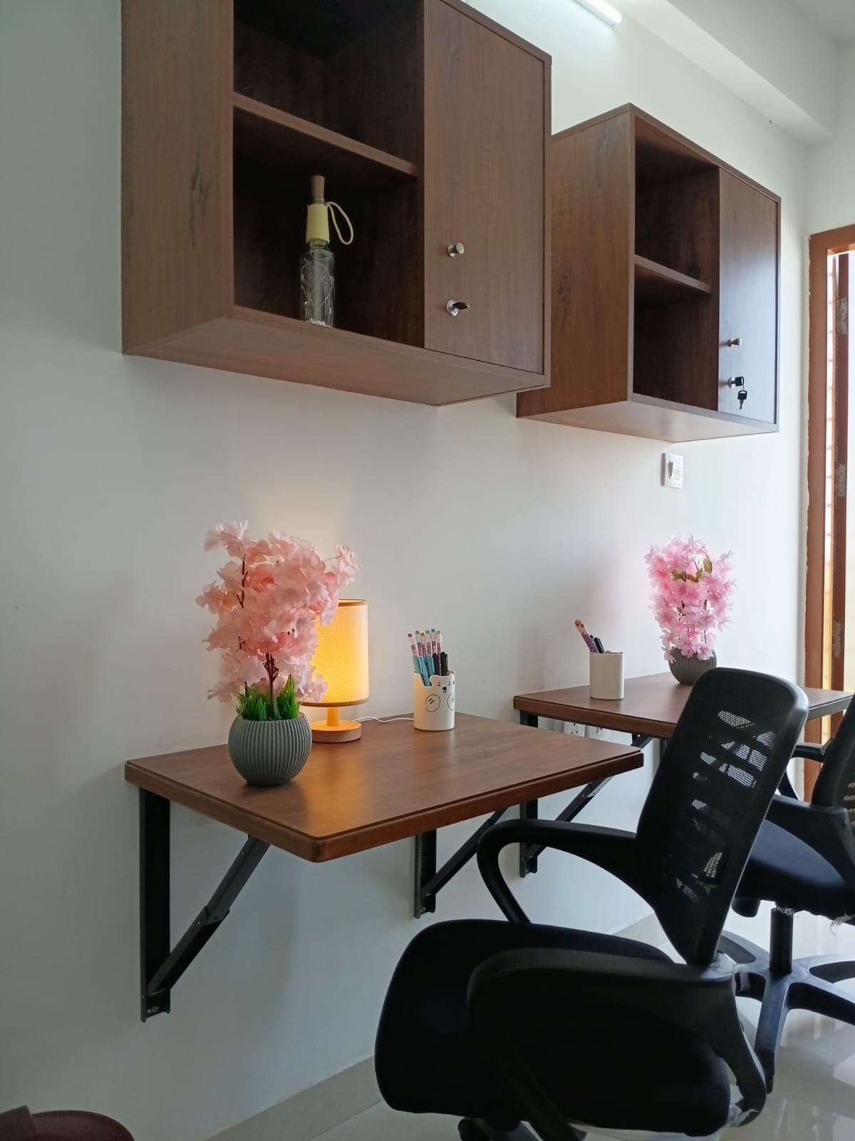 Two wall-mounted wooden desks with black mesh chairs, pink flowers, and overhead storage cabinets.