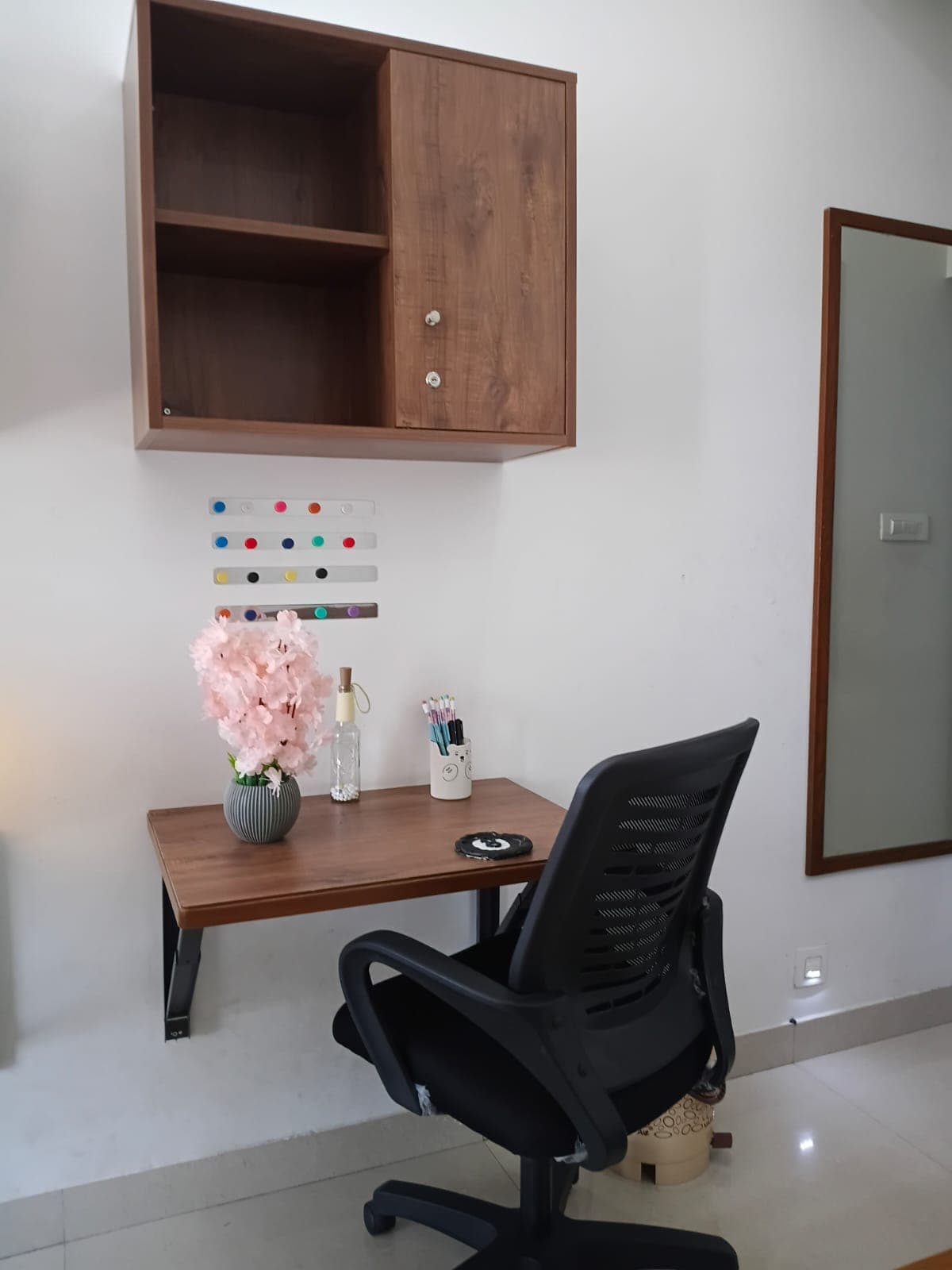 Wall-mounted wooden desk and cabinet with black office chair, pink flowers, and colorful magnets.